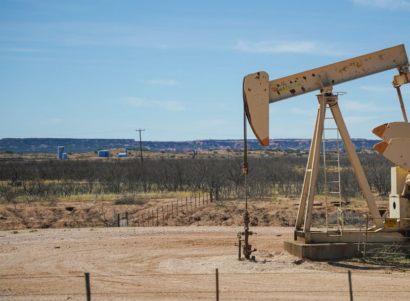 Gas well in dirt during day. Mountains and small trees in the background